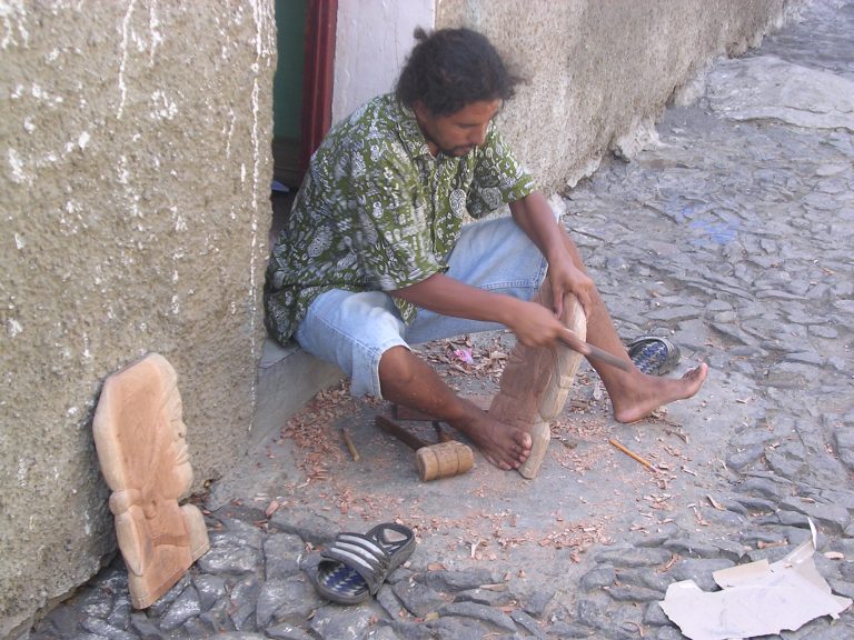 A barefoot Tony sitting on his stoop carving wood.