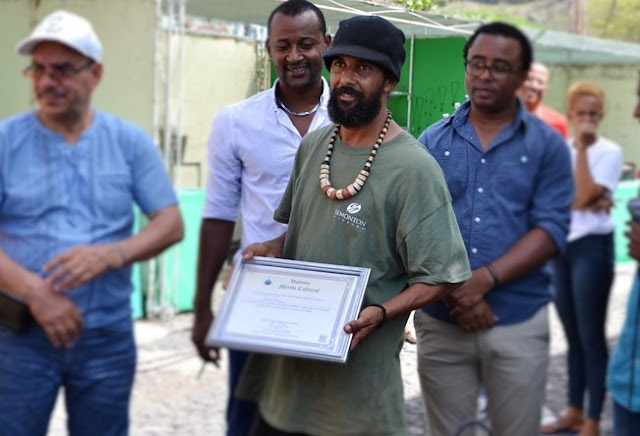 Several people, all African, surround Tony, a bearded man with a necklace, who is accepting an award.